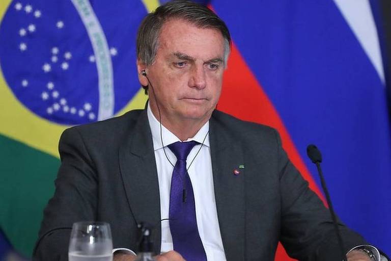 Imagem em primeiro plano mostra Jair Bolsonaro sério​, sentado de frente para uma mesa com um microfone. Ele usa fone de ouvido. Ao fundo, há uma bandeira do Brasil.