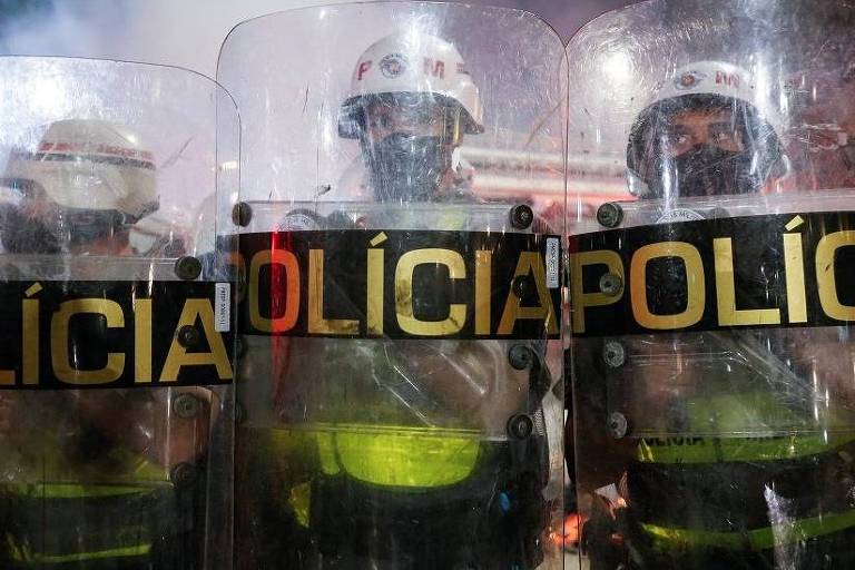 Três policiais seguram escudos. Eles estão um ao lado do outro e usam máscara.