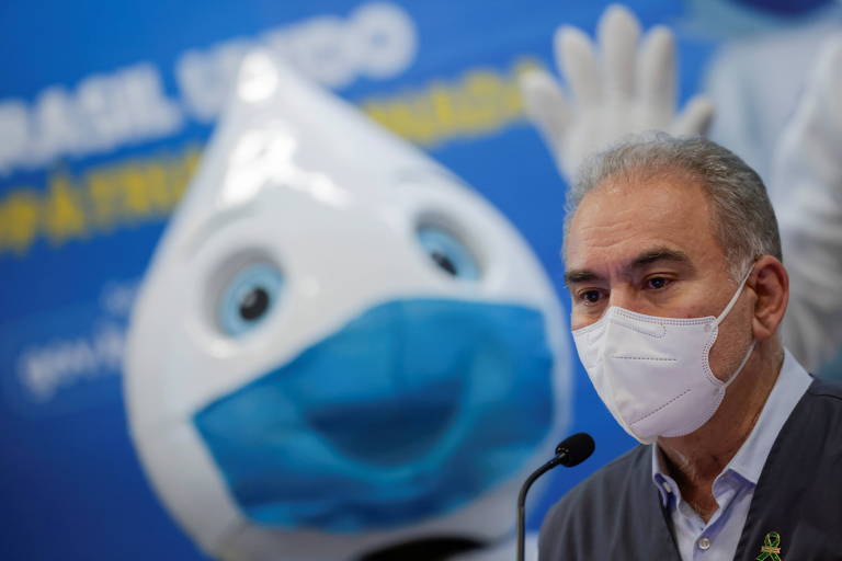 Queiroga usa máscara branca enquanto fala ao microfone; ao fundo, em um cartaz, há uma imagem do personagem Zé Gotinha usando também uma máscara, azul