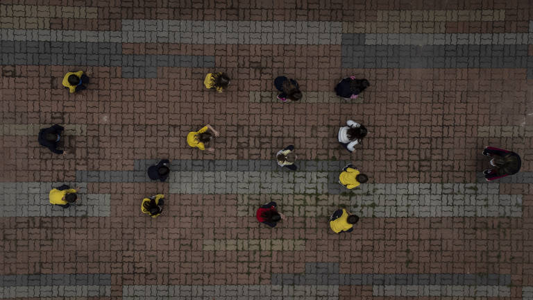 Quatorze crianças estão sentadas em um chão vermelho. Elas se organizaram em uma fileira com três pessoas, outra com quatro, mais uma com três e novamente uma com quadro delas. Na frente do grupo está uma mulher de cabelo preto em pé, ela é a professora do grupo. A foto foi tirada de cima.
