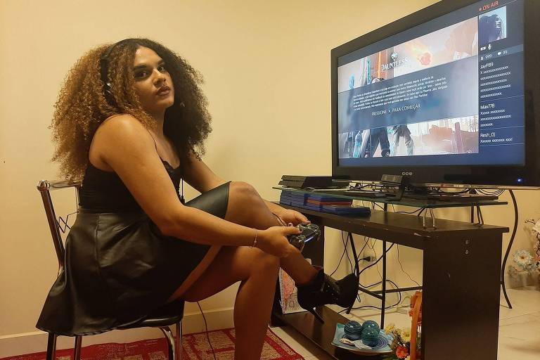 Mulher sentada de lado, com cabelos enrolados soltos, na cor castanhos claros, na altura do peito, de pernas cruzadas,  de saia e regata preta, segurando um controle de videogame preto, em frente a uma televisão ligada