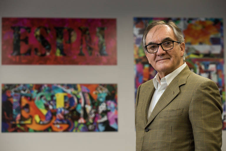 Homem branco grisalho de óculos à frente de uma parede com painéis coloridos