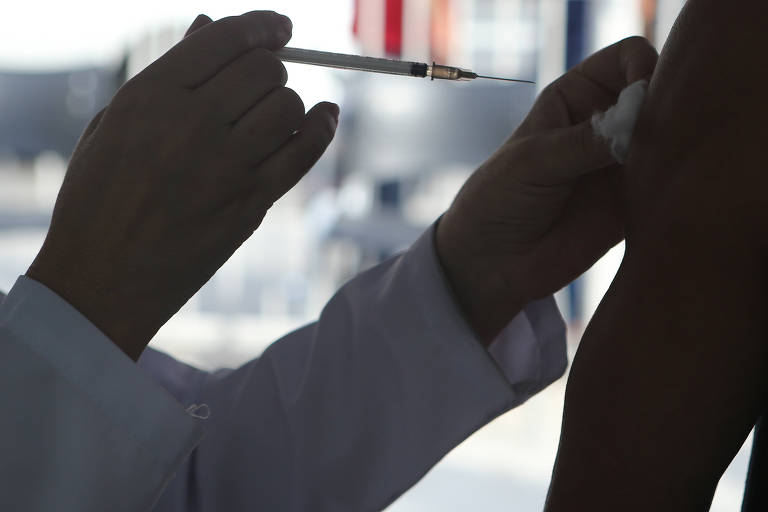 Mãos de pessoa com avental branco de manga longa aplicam vacina em braço. Imagem contra a luz