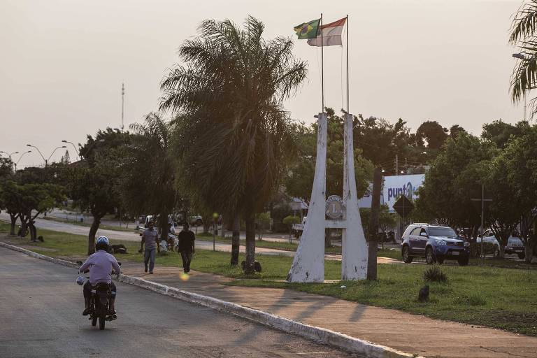 Imagem mostra avenida que divide as cidades de Capitan Bado (Paraguai) e Coronel Sapucaia (Brasil). Na via, há uma motocicleta e em uma calçada pedestres. Ao fundo, as bandeiras dos dois países estão hasteadas.