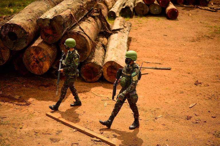 Dois soldados usando roupa verde camuflada e segurando armas caminham ao lado de troncos de árvore empilhados no chão