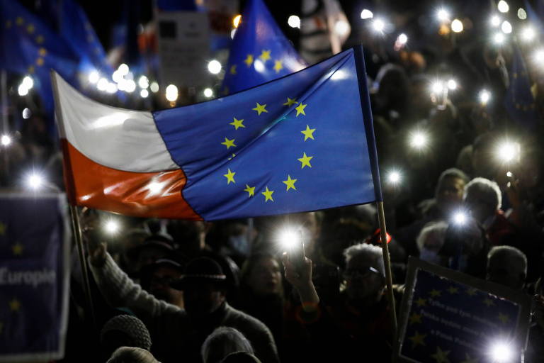 Várias pessoas seguram celular aceso durante à noite em manifestação onde pessoas levam bandeira branca e vermelha, da polônia, e azul com círculo de estrelas amarelas, da união europeia