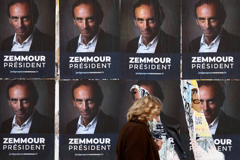 Uma mulher de máscara no rosto anda em frente a uma parede com vários cartazes com o rosto do polemista de extrema-direita Eric Zemmour. É possível ler a frase "Zemmour President", ou "Zemmour Presidente" em português escrita abaixo da foto dele nos cartazes