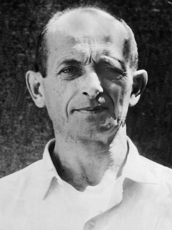 Foto antiga, em preto e branco, de Adolf Eichmann, vestindo uma camisa de botão. Ele é um homem careca, com cabelos apenas nas laterais da cabeça e tem a testa franzida