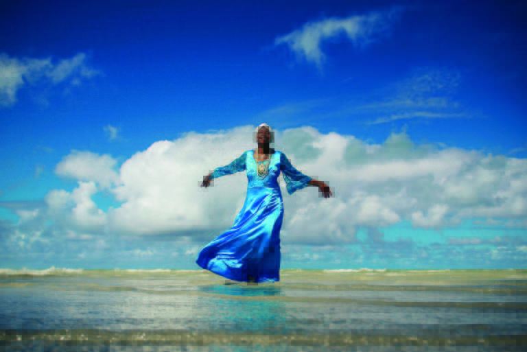 Uma mulher negra está em pé de braços abertos, vestindo um vestido azul, na frente do mar