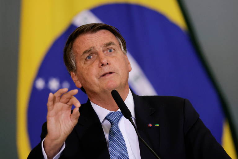 Bolsonaro, um homem branco e de terno, gesticulando com uma das mãos enquanto discursa. Ao fundo, uma bandeira do Brasil