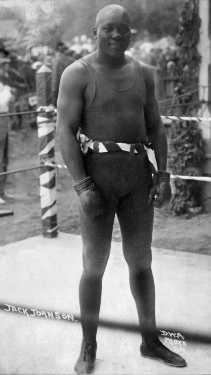A imagem apresenta o boxeador Jack Johnson, homem negro, forte, vestindo calção e luvas de boxe, além de uma botina esportiva com cadarços.