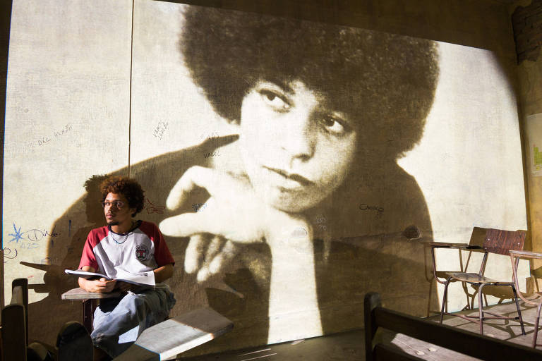 Em destaque, imagem antiga de ativista Angela Davis é projetada em uma parede. No canto esquerdo da foto, um garoto está sentada em uma carteira escolar