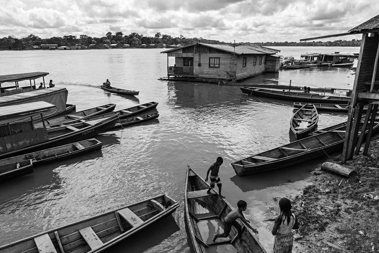 Crianças brincando à beira de um rio no Amazonas, em meio a pequenos barcos de madeira, com casas erguidas sobre as águas e a outra margem do rio à distância.  