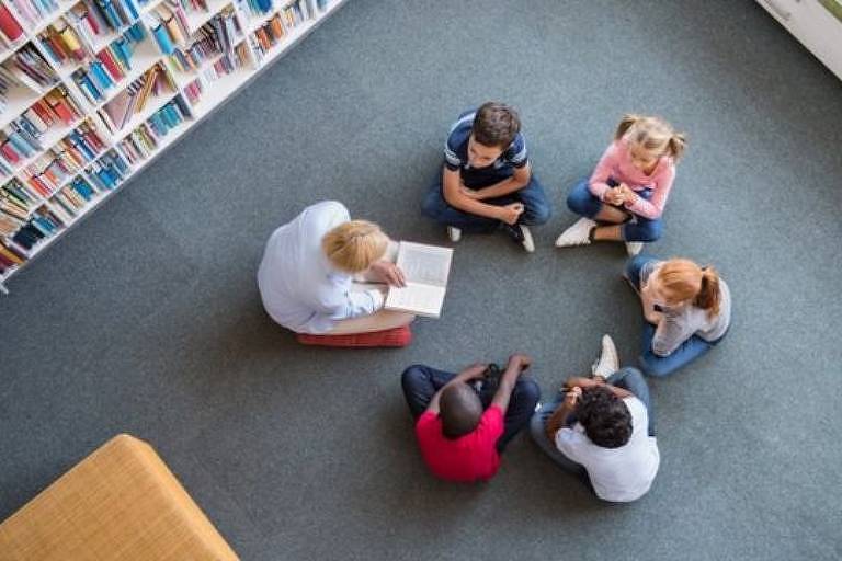 Imagem aérea mostra um grupo de cinco crianças e uma adulta sentados no chão formando um círculo. As crianças observam a adulta, que está com um livro na mão. No canto esquerdo da imagem, há uma estande com livros