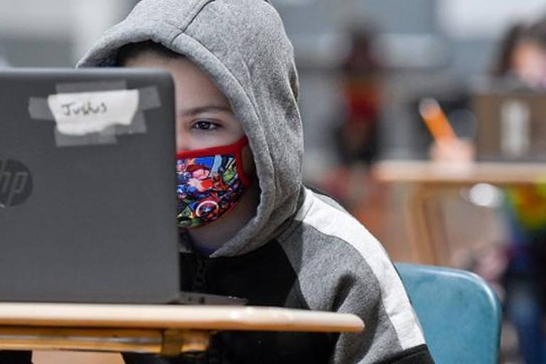Imagem em primeiro plano mostra uma criança de máscara e blosa de capuz sentada em uma carteira escolar. Ela olha para a tela do computador sobre a mesa
