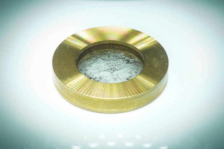 Círculo dourado com algumas pedrinhas cor de cinza no meio, protegidas por um vidro. Uma luz forte ilumina o objeto