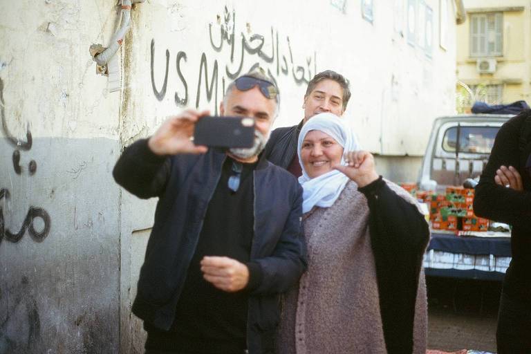 Karim Aïnouz está em pé segurando o celular para tirar uma selfie ao lado de uma mulher com um lenço cobrindo sua cabeça 