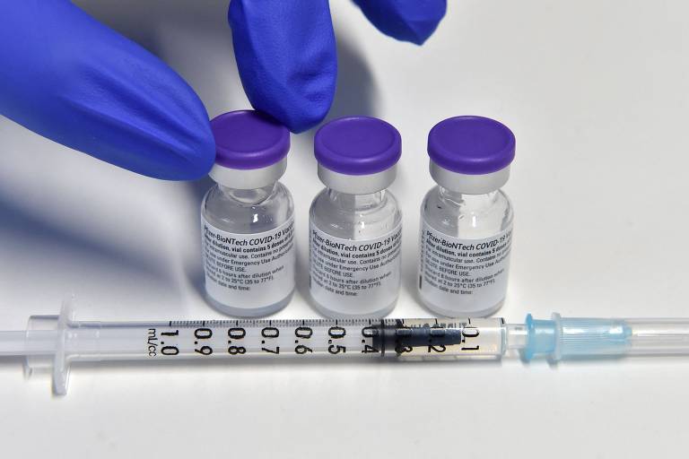 Três frascos de vacina da Pfizer dispostos sobre uma mesa e uma seringa. Um mão com luva apoia os dedos em um dos recipientes