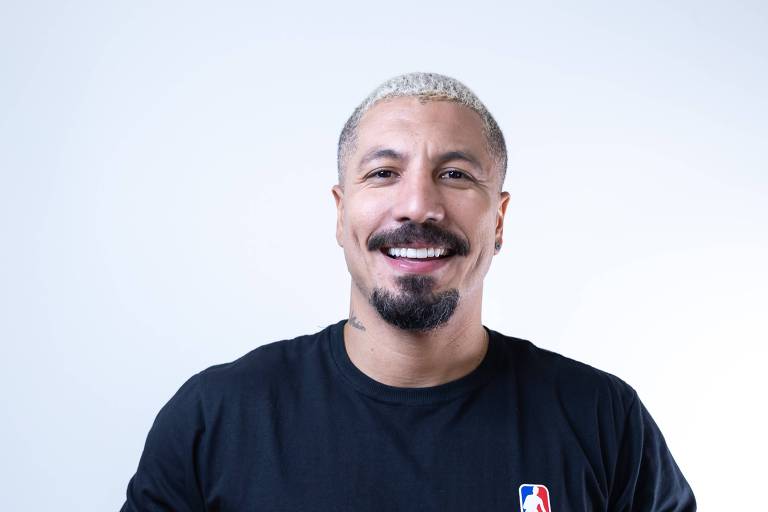 Homem careca com camiseta preta com símbolo da NBA segurando bola de basquete sorrindo em frente a um fundo branco