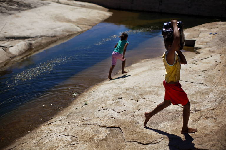 Duas crianças carregam água de rio que passa entre terra seca