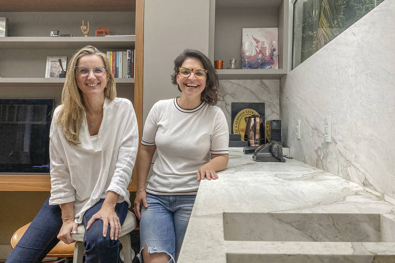 Duas mulheres brancas sentadas em uma casa com uma pia ao lado. Ambas usam blusa branca e calças jeans