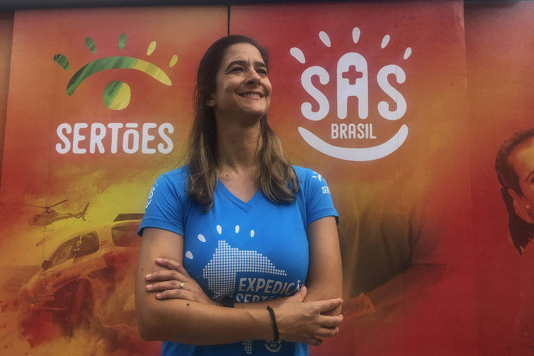 mulher loira de blusa azul cruza os braços sorrindo em frente a painé laranja escrito SAS Brasil e Sertões