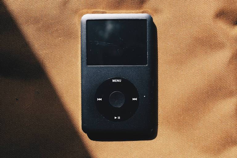 Foto mostra iPod preto em cima de pano marrom claro no sol