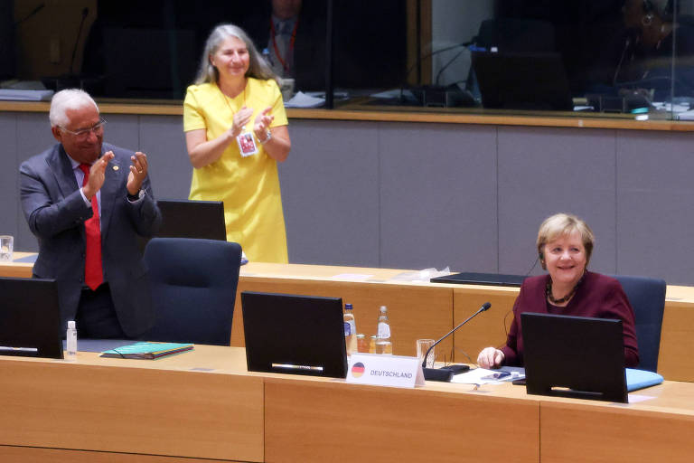 Merkel, de roupa vinho, sentada, sorri enquatno é aplaudida por homem de pé, de terno, gravata vermelha e máscara contra covid
