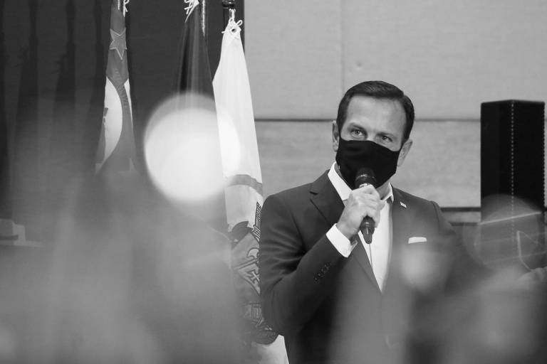 Homem branco de cabelos pretos curtos, com terno e gravata e máscara de proteção contra Covid escura, segurando microfone para falar em evento. Ao seu lado, há algumas bandeiras. 