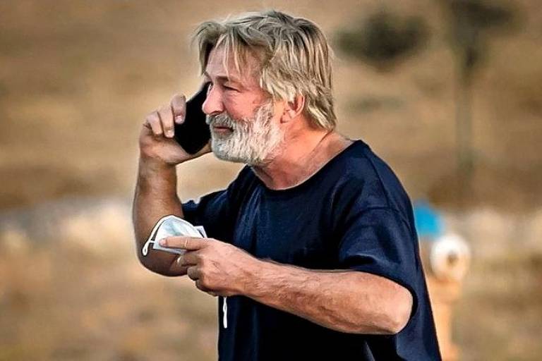 Em foto colorida, homem de camisa preta fala ao telefone enquanto segura a máscara de proteção em uma das mãos