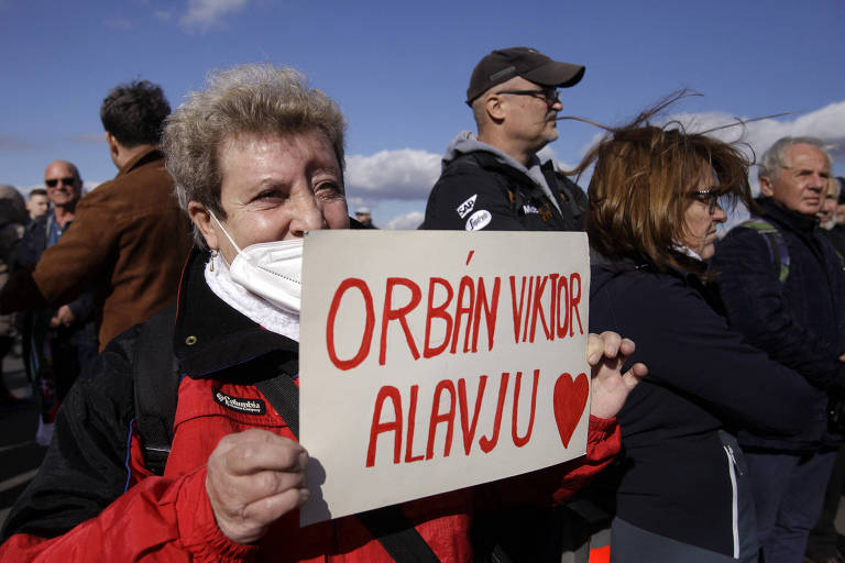Idosa de casaco vermelho segura cartaz onde está escrito "Orbán Viktor A Lav Ju", ou seja, Orbán eu te amo, em letras vermelhas