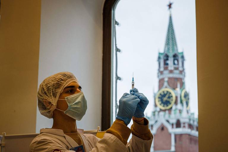 Profissional de saúde veste toucas, jaleco e luvas, e segura uma seringa de vacina; ao fundo, um prédio típido da Rússia