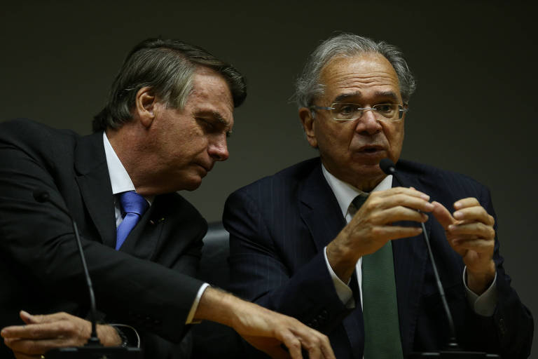 Bolsonaro e Guedes são homens idosos, de cabelos grisalhos, que vestes ternos pretos e estão lado a lado