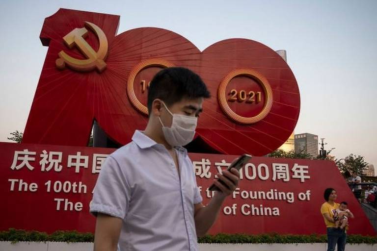 homem chinês de máscara caminha com celular na mão em frente a monumento em vermelho