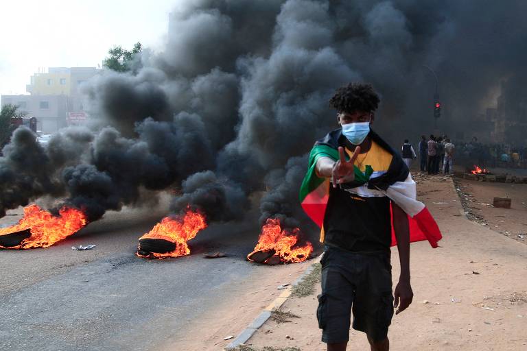 Manifestante sudanês coberto com a bandeira nacional ao lado de pneus em chamas durante uma manifestação na capital, Cartum; pessoas saíram às ruas após ter início um golpe militar no país da África Oriental: o premiê e diversas figuras civis do governo foram detidos ilegalmente 