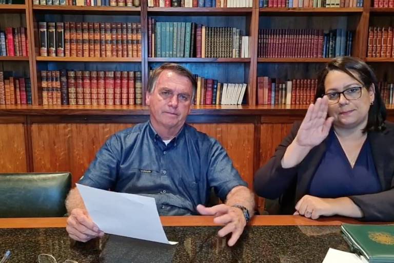 Bolsonaro, de camisa azul, sentado ao lado de uma intérprete de libras que veste preto
