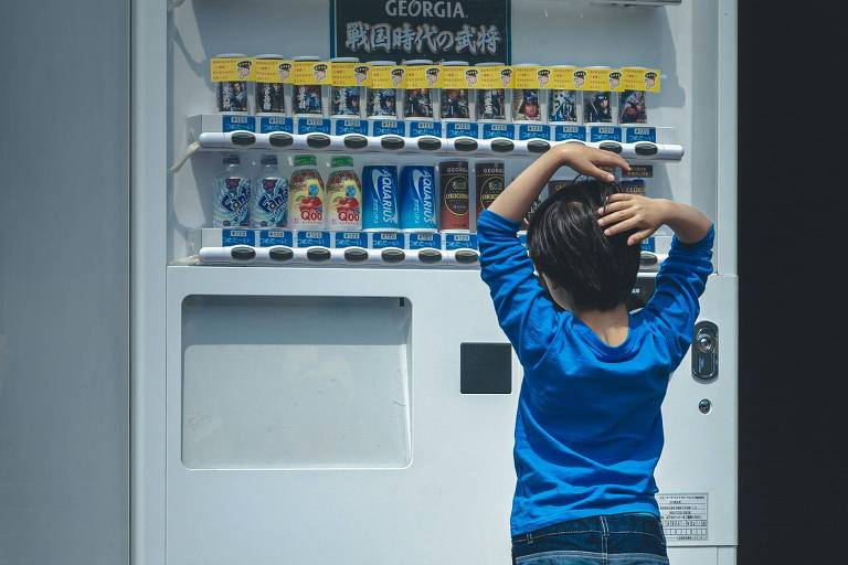 Foto mostra criança de costas olhando para uma prateleira com vários tipos de bebida à venda. Ela veste uma camiseta azul e tem os braços sob a cabeça, como se estivesse em dúvida do que escolher.