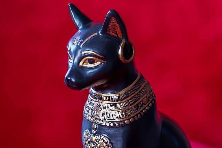 Foto mostra estátua de gato preto com adereços dourados, próprios da cultura egípcia. A estátua está sob um tecido vermelho que também cobre o fundo da foto.
