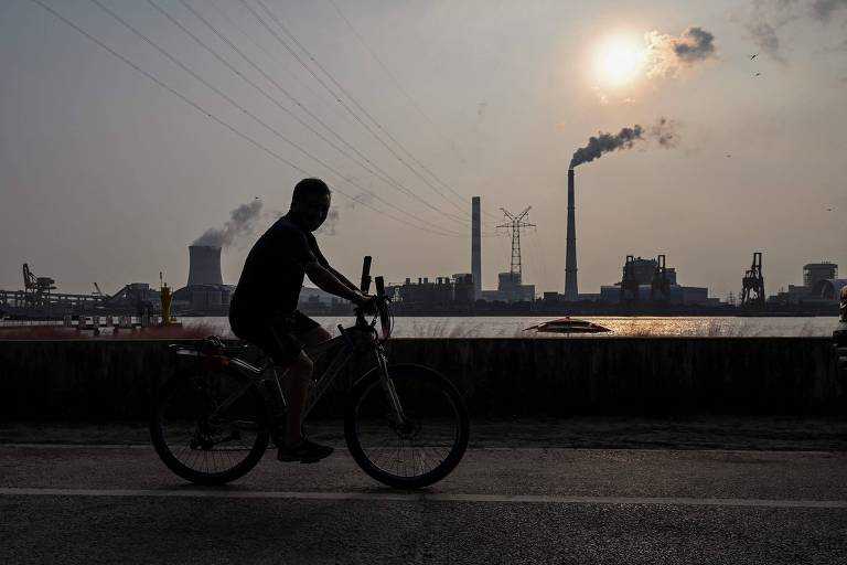 Em destaque, homem anda de bicicleta às margens do um rio. Ao fundo, se vê uma estação de energia soltando fumaça no ar