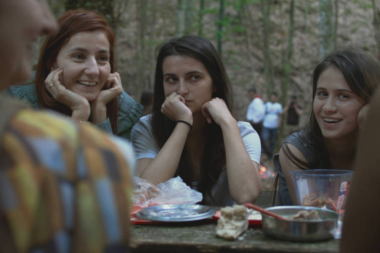 Três mulheres sentadas em uma mesa numa floresta. Eles estão com a mão apoiada no rosto