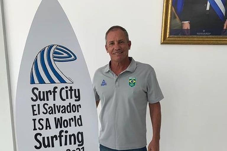 Presidente da Confederação Brasileira de Surf (CBSurf), Adalvo Argolo, ao lado de placa do ISA Games, torneio de surfe, que em 2021 foi disputado em El Salvador