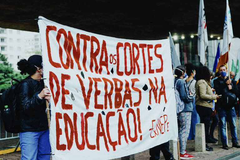 Pessoas em protesto seguram cartaz com letras vermelhas contra cortes na educação