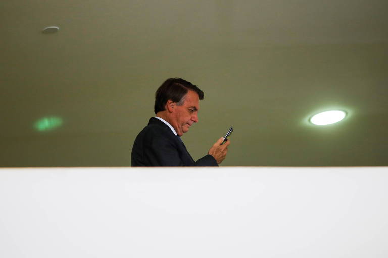 Bolsonaro, um homem branco, de cabelos e terno escuros, aparece da altura do peito pra cima, por trás de um muro branco, mexendo no celular 