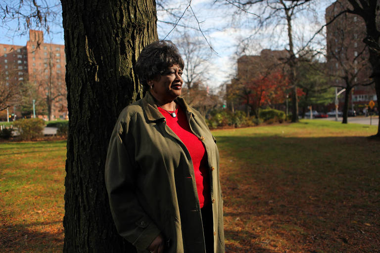 A ativista americana Claudette Colvin na frente de uma árvore; ela é uma mulher negra e usa uma blusa vermelha com um casaco verde longo