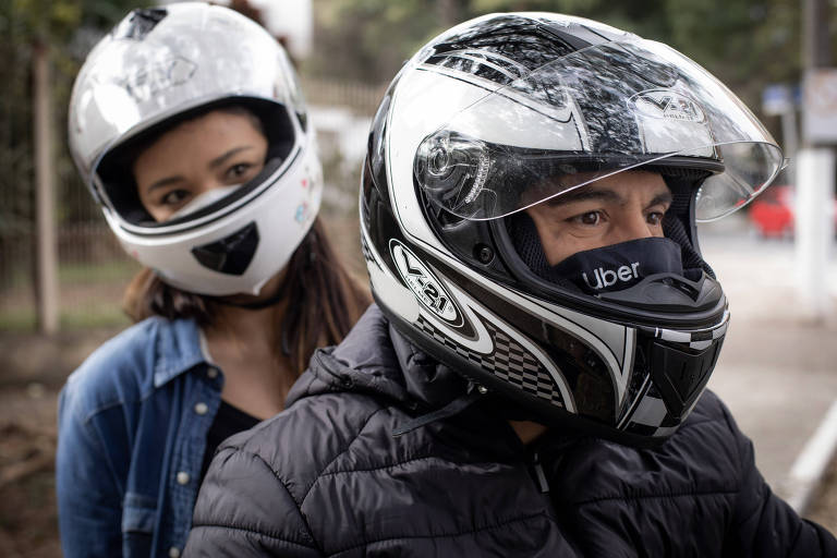 A foto mostra um homem e uma mulher em uma moto utilizando capacetes, o homem está na frente e utiliza uma máscara com a inscrição Uber