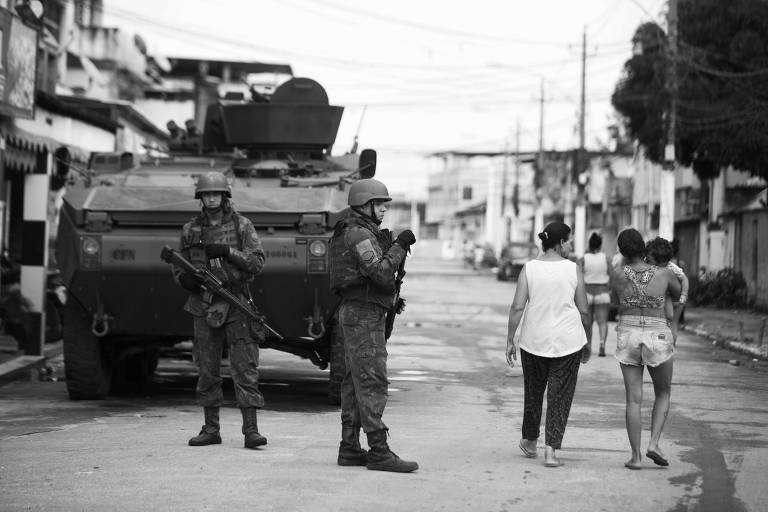 Dois soldados de uniforme e capacete, segurando fuzis, na frente de um blindado, observam o movimento numa rua asfaltada, enquanto duas moradoras do local passam por eles. Uma das mulheres tem uma criança no colo.