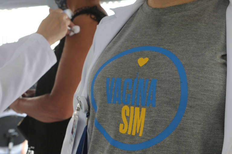 Em primeiro plano, uma pessoa com uma camiseta onde se lê "Vacina sim"; ao fundo, uma pessoa é vacinada 