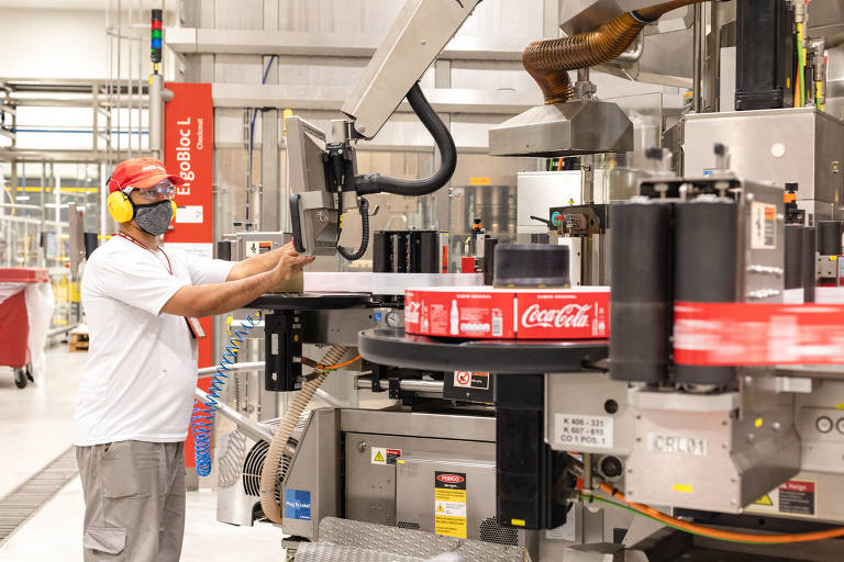 Homem aparece com protetores de ouvido, chapéu da Coca-Cola, máscara e óculos operando uma máquina. No equipamento, é possível ver rótulos de garrafas da Coca-Cola.