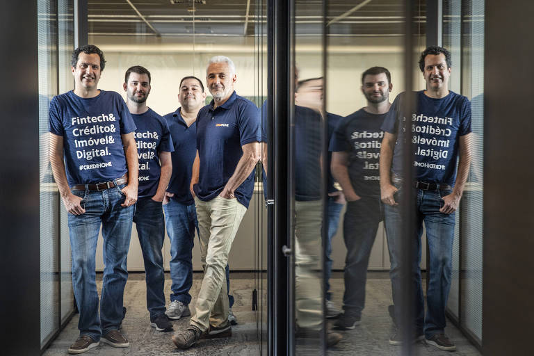 Quatro homens brancos com camisetas azuis, em pé, em corredor com paredes de vidro 