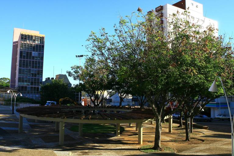 Na foto, é possível ver a praça, com várias árvores altas fazendo sombra numa estrutura circular de concreto. No fundo, prédios altos do centro de São Carlos. Não aparecem pessoas na praça